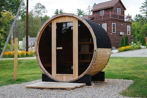 Una gran estructura de barril de madera con una puerta, ubicada en una zona de grava con una casa al fondo.