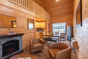 Interior de una cabaña de madera con chimenea, sillones de cuero, una mesa de comedor, una zona de cocina y un balcón.