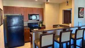 A modern kitchen with wooden cabinets, a black refrigerator, a microwave, and a stove. There are three bar stools with cushioned seats.