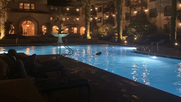 A nighttime pool area with lounge chairs and palm trees, surrounded by a building with illuminated windows.