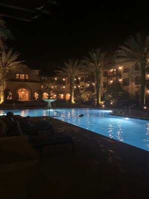 A nighttime pool area with lounge chairs and palm trees, surrounded by a building with illuminated windows.