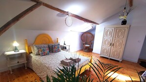 A bedroom with a sloped ceiling, a bed with patterned bedding, a nightstand with a lamp, a wicker chair, a cabinet, and a potted plant. - Aluguer apartamento Gaivota (Portimão)