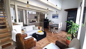 A spacious living area with a wooden floor, a sofa, two armchairs, a dining table with chairs, a staircase, and a kitchen in the background. - Aluguer apartamento Gaivota (Portimão)