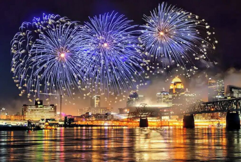 A city skyline with fireworks over a body of water, reflecting city lights.