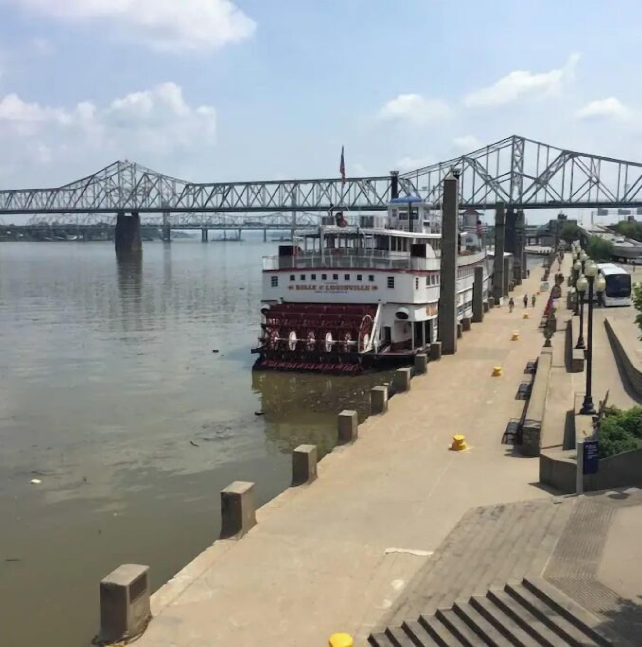 A riverfront with a large white boat docked at a pier, a bridge in the background, and a sidewalk with bollards and street lamps.