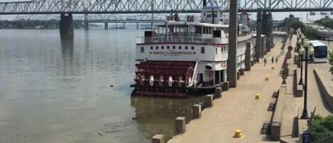 A riverfront with a large white boat docked at a pier, a bridge in the background, and a sidewalk with bollards and street lamps.