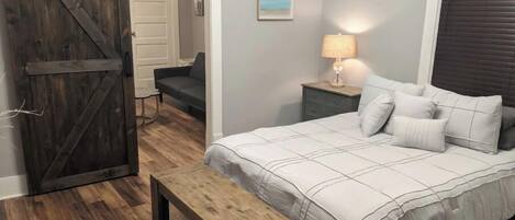 A bedroom with a bed, a bench, a nightstand, a lamp, a door, and a painting on the wall.