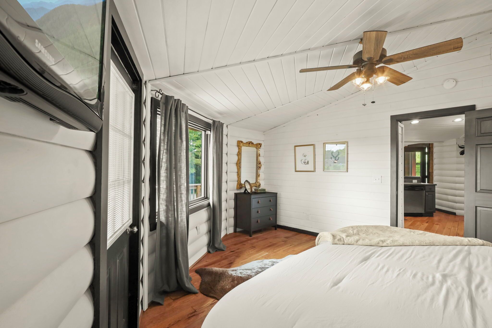 Quarto com ventoinha de teto em madeira, cama com edredom branco, cômoda e espelho na parede.
