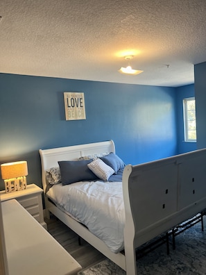 A bedroom with a bed, a nightstand, a lamp, a wall sign, and a window.