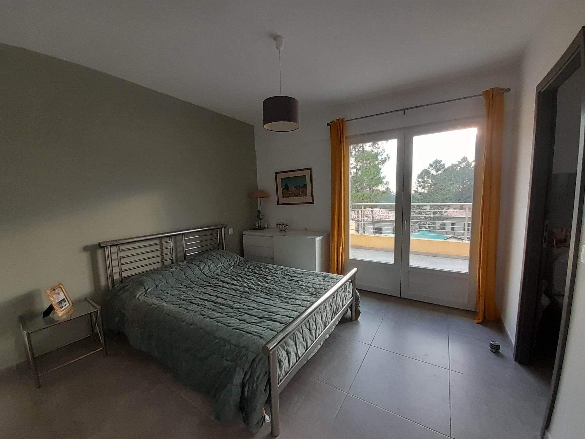 A bedroom with a bed, a bedside table, a lamp, a picture frame, a window with curtains, and a view of a swimming pool.