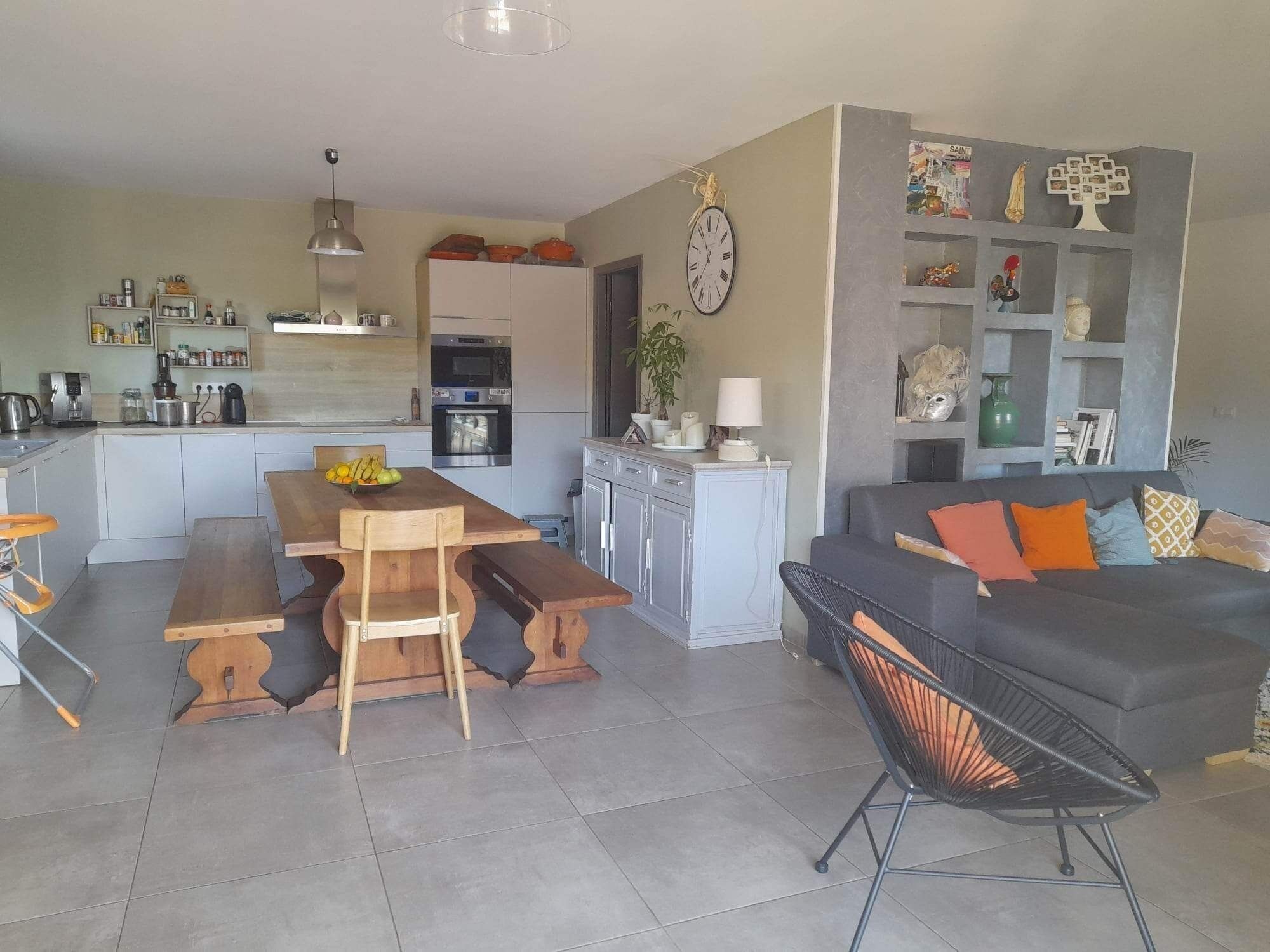 A modern kitchen with a central island, a dining area with a wooden table and chairs, a grey sofa with colorful cushions, and a black chair with a woven backrest.