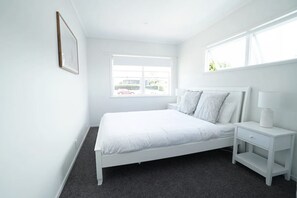 A white bedroom with a bed, nightstand, and window. - Bright & Central Home with Mini Golf Area (Auckland)