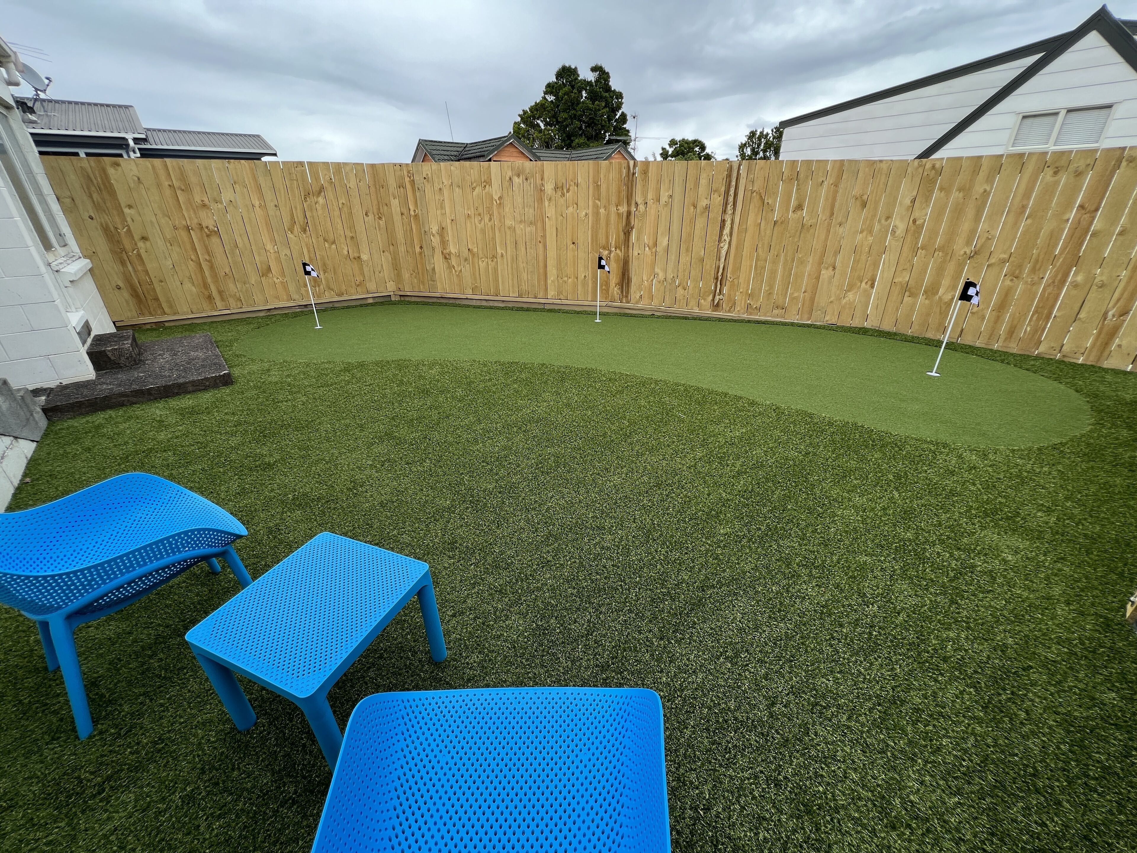 Un jardin avec une pelouse artificielle, une clôture en bois et deux chaises en plastique bleues.