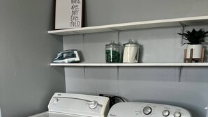 A laundry room with a front-loading washer and dryer, a shelf with jars and a plant, and a sign above the shelf. - 600 House (West Lafayette)