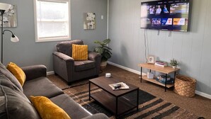 A living room with a grey couch, a coffee table, a TV mounted on the wall, and a window with blinds. - 600 House (West Lafayette)