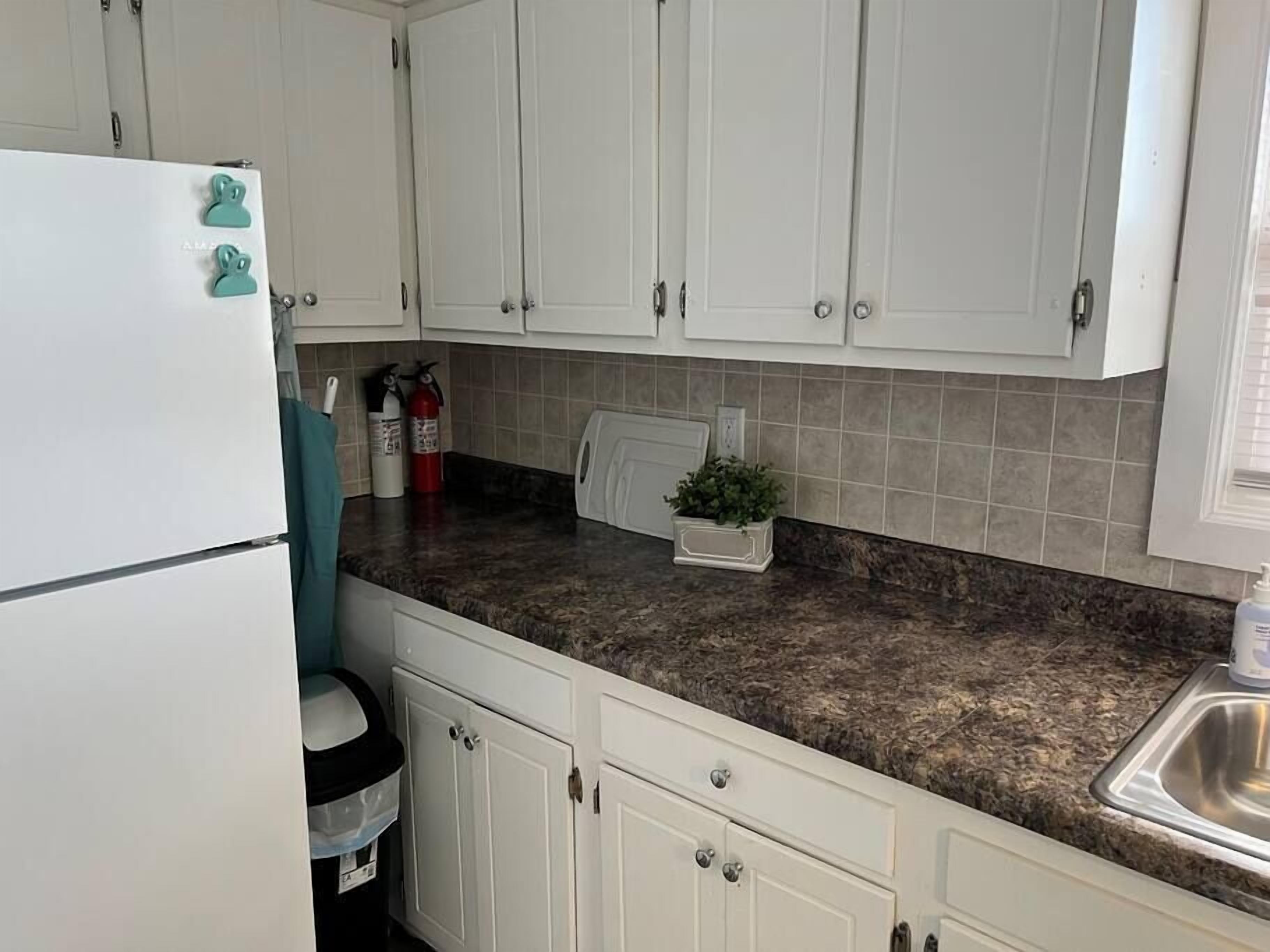 A kitchen with white cabinets, a granite countertop, a stainless steel sink, a refrigerator, and a fire extinguisher.