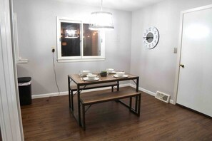 A dining area with a wooden table set for four, a wall clock, and a window with a hanging light fixture. - Salisbury (West Lafayette)