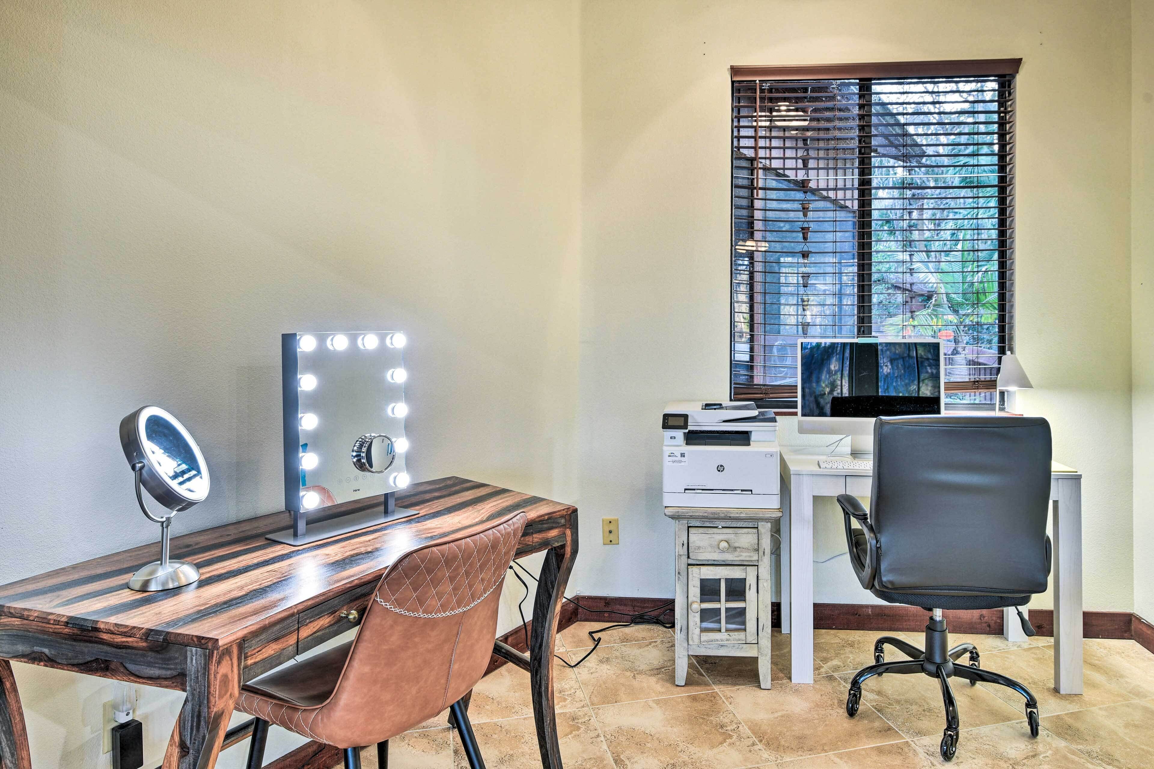 A room with a wooden desk, a mirror with lights, a black office chair, a printer, and a window with blinds.