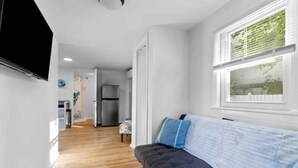 A modern living room with a grey sofa, a flat-screen TV mounted on the wall, and a large window with blinds. - Cottage by the Sea (Warwick)