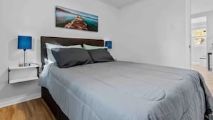 A bedroom with a bed, a nightstand, a lamp, and a painting on the wall. - Cottage by the Sea (Warwick)