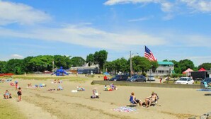 A beach with people sunbathing, a large American flag, and a building with a green roof in the background. - Cottage by the Sea (Warwick)