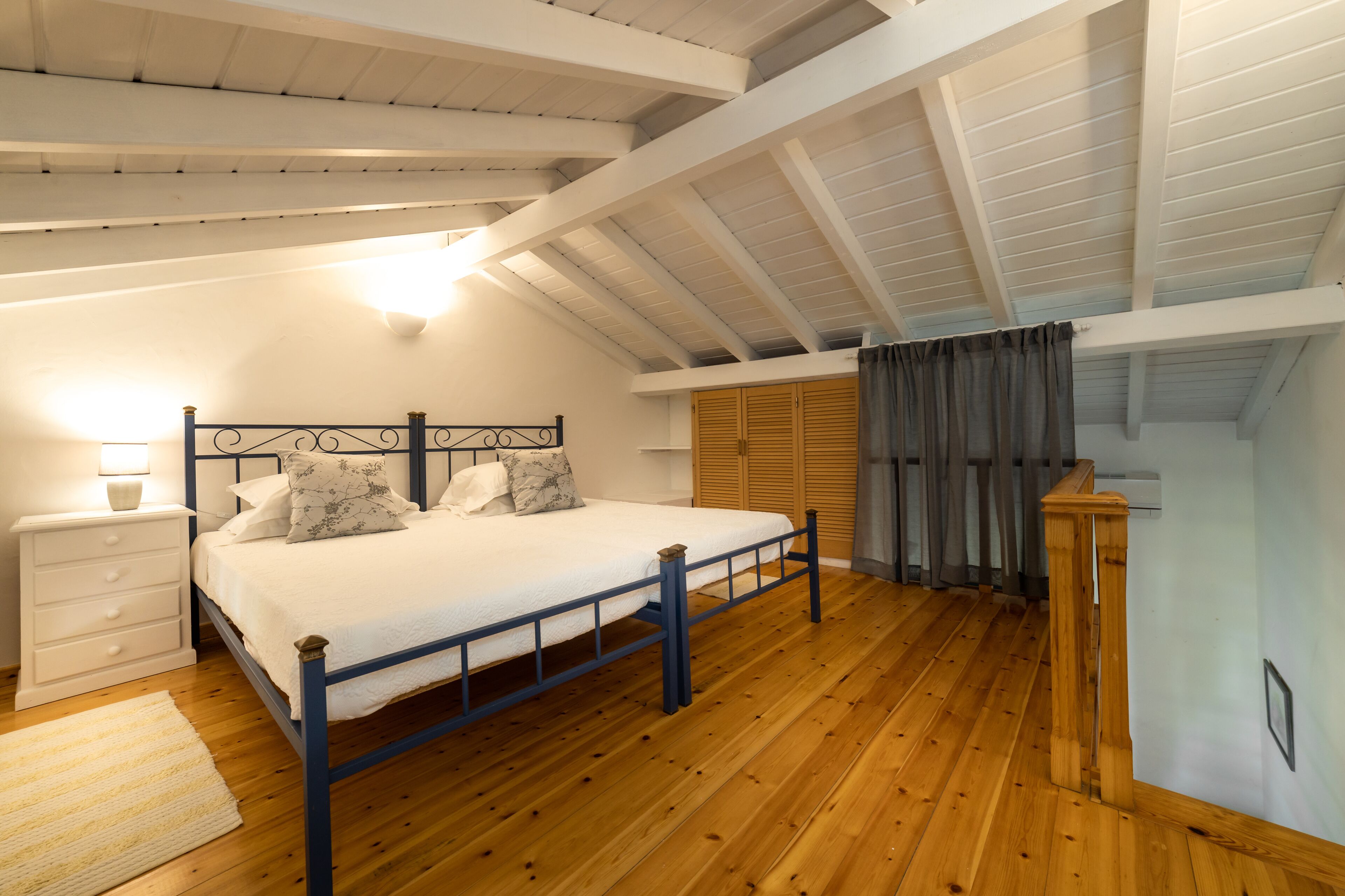 Un dormitorio con suelo de madera, una cama con estructura metálica, una mesita de noche, una lámpara y una ventana con cortinas.