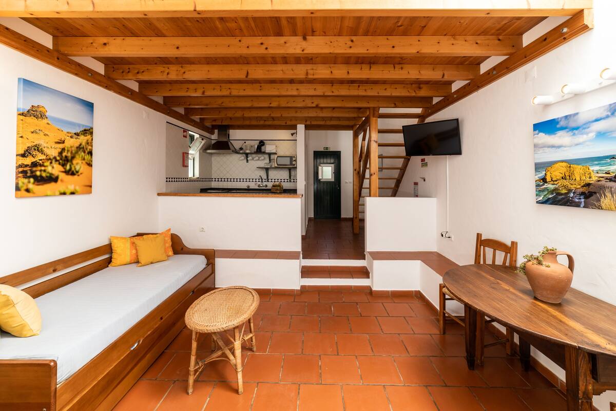 A cozy room with a wooden ceiling, a bed with yellow pillows, a dining table with chairs, and a TV mounted on the wall.