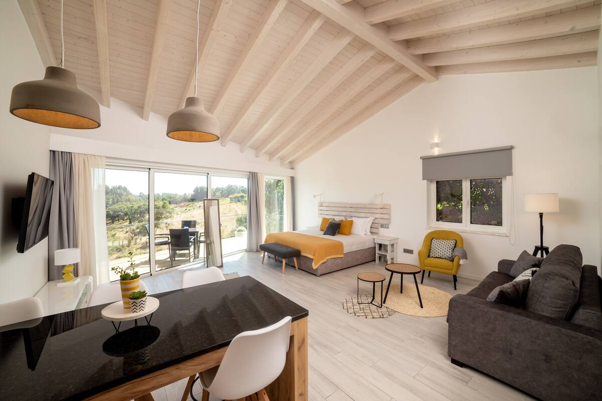 A spacious room with a bed, a sofa, a dining area, and a large window offering a view of the outdoors.