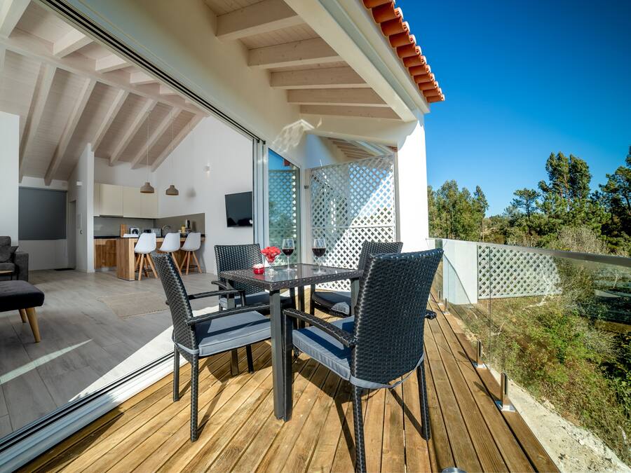 Una terraza de madera con mesa y sillas, una zona de cocina y vistas a árboles.