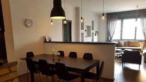 Un comedor moderno con una mesa grande y sillas, una lámpara colgante y vistas a una sala de estar con sofá y cortinas.