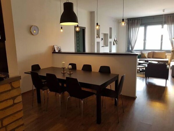 Ein moderner Essbereich mit einem großen Tisch und Stühlen, einer Hängelampe und Blick auf eine Sitzecke mit Sofa und Vorhängen.