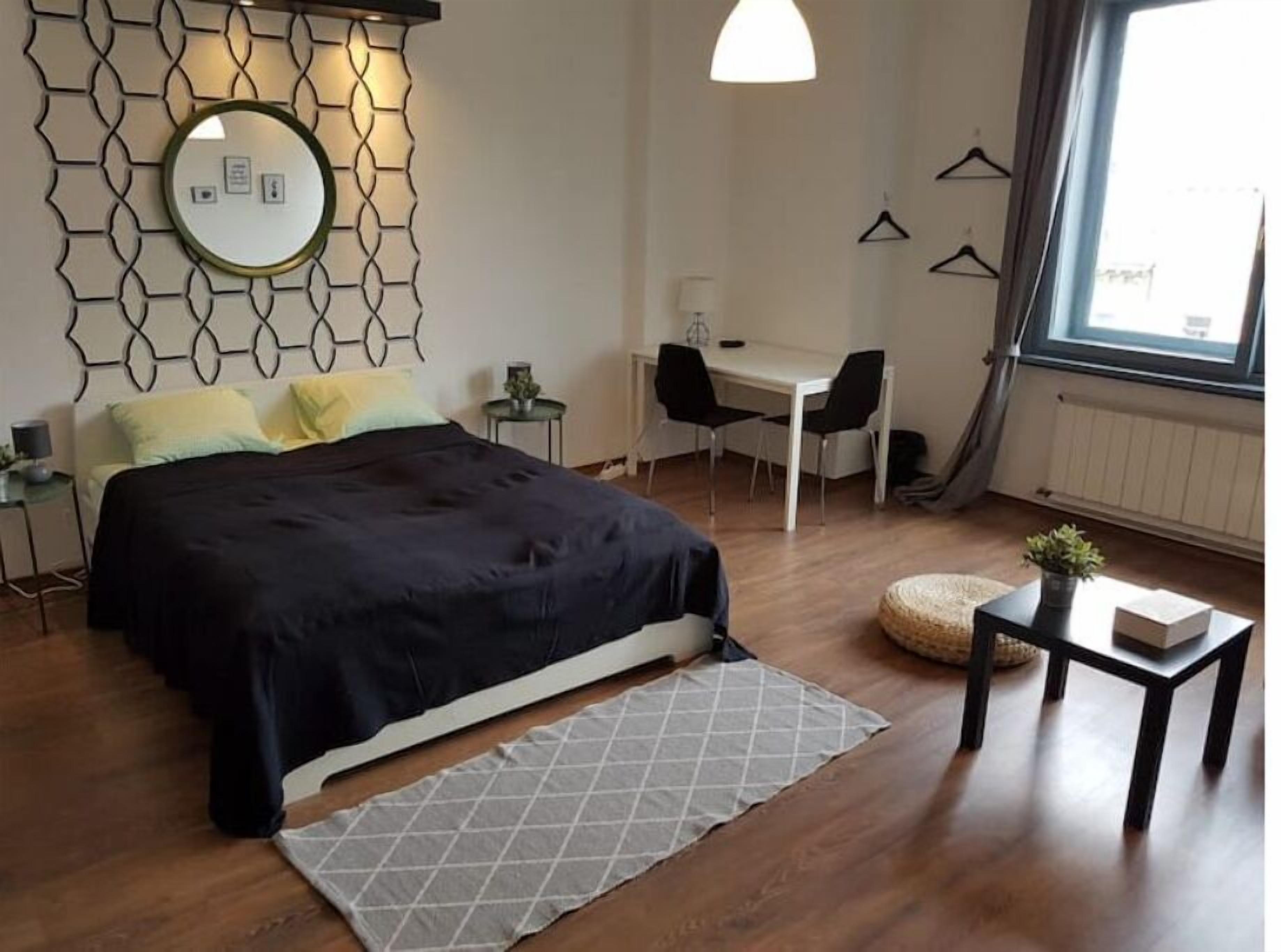 Una camera da letto moderna con un letto, una scrivania e una sedia. Si vede una finestra con tende, uno specchio fissato alla parete e una lampada a sospensione.