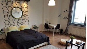 Un dormitorio moderno con cama, escritorio y silla. Hay una ventana con cortinas, un espejo montado en la pared y una lámpara colgante.