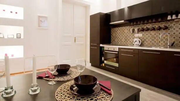 A modern kitchen with dark cabinets, a countertop, and a backsplash. There is a dining table with a placemat, a candle, and a wine glass. - Downtown Rising Sun (104sqm 2bedr/2bath/living) (Budapest)