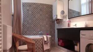 A bathroom with a bathtub, a sink, a mirror, a washing machine, and a wicker chair. - Downtown Rising Sun (104sqm 2bedr/2bath/living) (Budapest)