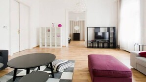 A modern living room with a sofa, ottoman, and a coffee table. There is a white shelving unit with decorative items and a television in the background. - Downtown Rising Sun (104sqm 2bedr/2bath/living) (Budapest)