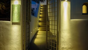 A well-lit staircase leading to a door with a decorative gate, surrounded by walls with light fixtures and a palm tree. - Oia Treasures Art Suites (Santorini)