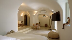 A spacious room with a large bed, a dining area, and a TV mounted on the wall. - Oia Treasures Art Suites (Santorini)