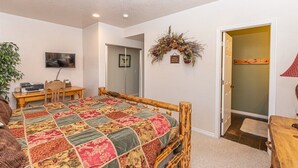 Un dormitorio con una cama de madera, un escritorio con silla, un televisor y una puerta que da a otra habitación.