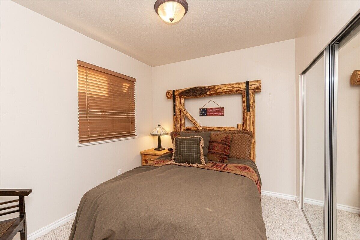 Ein Schlafzimmer mit einem Bett, einem Nachttisch, einer Lampe, einem Fenster mit Jalousien und einem Holzkopfstück.
