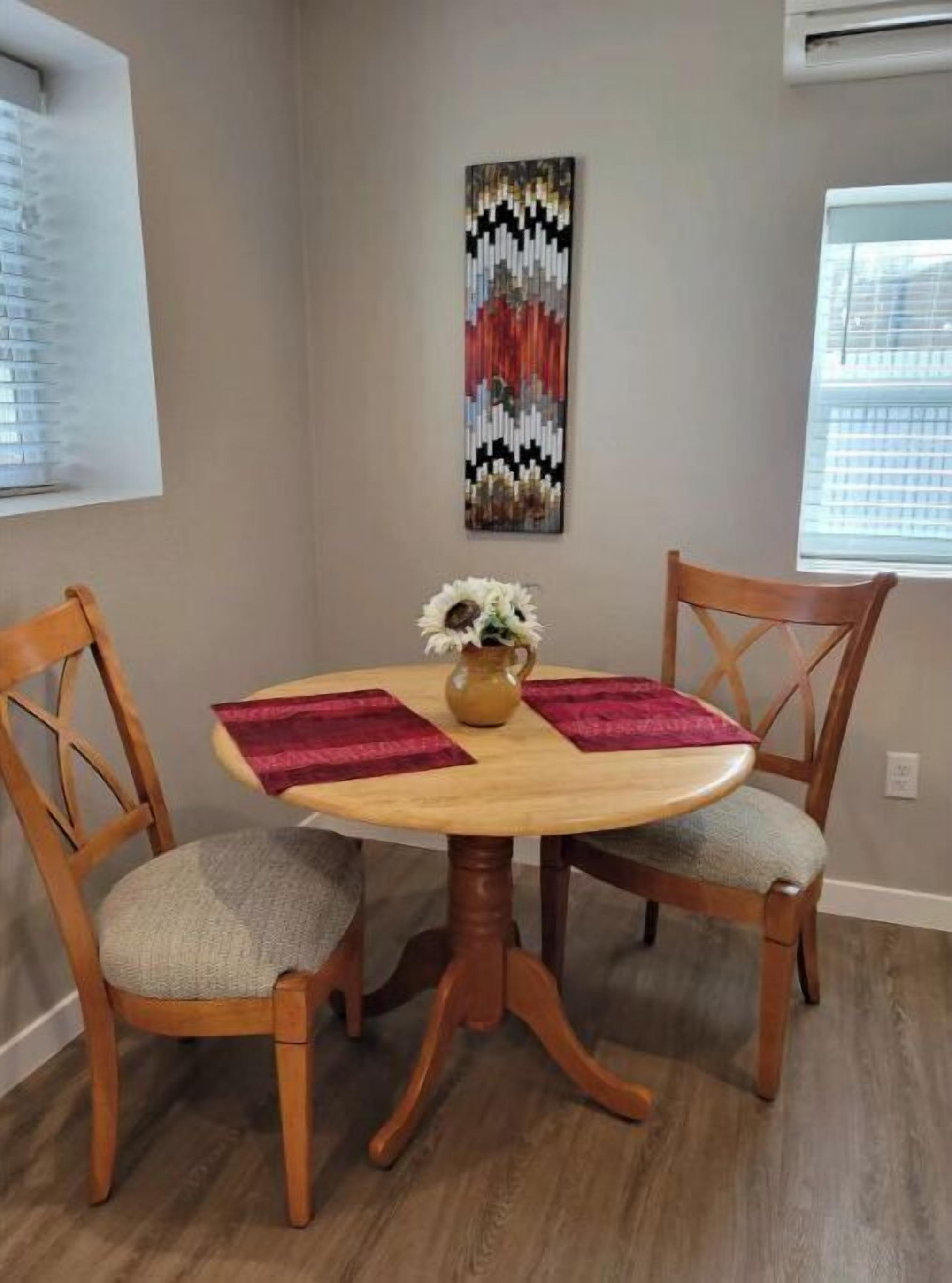 A dining area with a round wooden table, two wooden chairs with cushioned seats, a vase with flowers, and a colorful abstract painting on the wall.