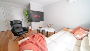 A modern living room with a white sofa, a black leather chair, a small round table with a vase, and a flat-screen TV mounted on the wall displaying 'NETFLIX'. - 4 Bedroom fun house - spa, sauna, fire pits, pool table, 4 TV's, 2 outdoor areas (Healesville)