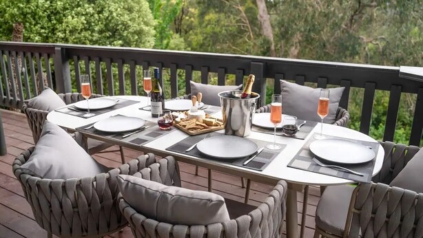 An outdoor dining area with a table set for four, surrounded by cushioned chairs, with a view of trees and a wooden deck. - 4 Bedroom fun house - spa, sauna, fire pits, pool table, 4 TV's, 2 outdoor areas (Healesville)