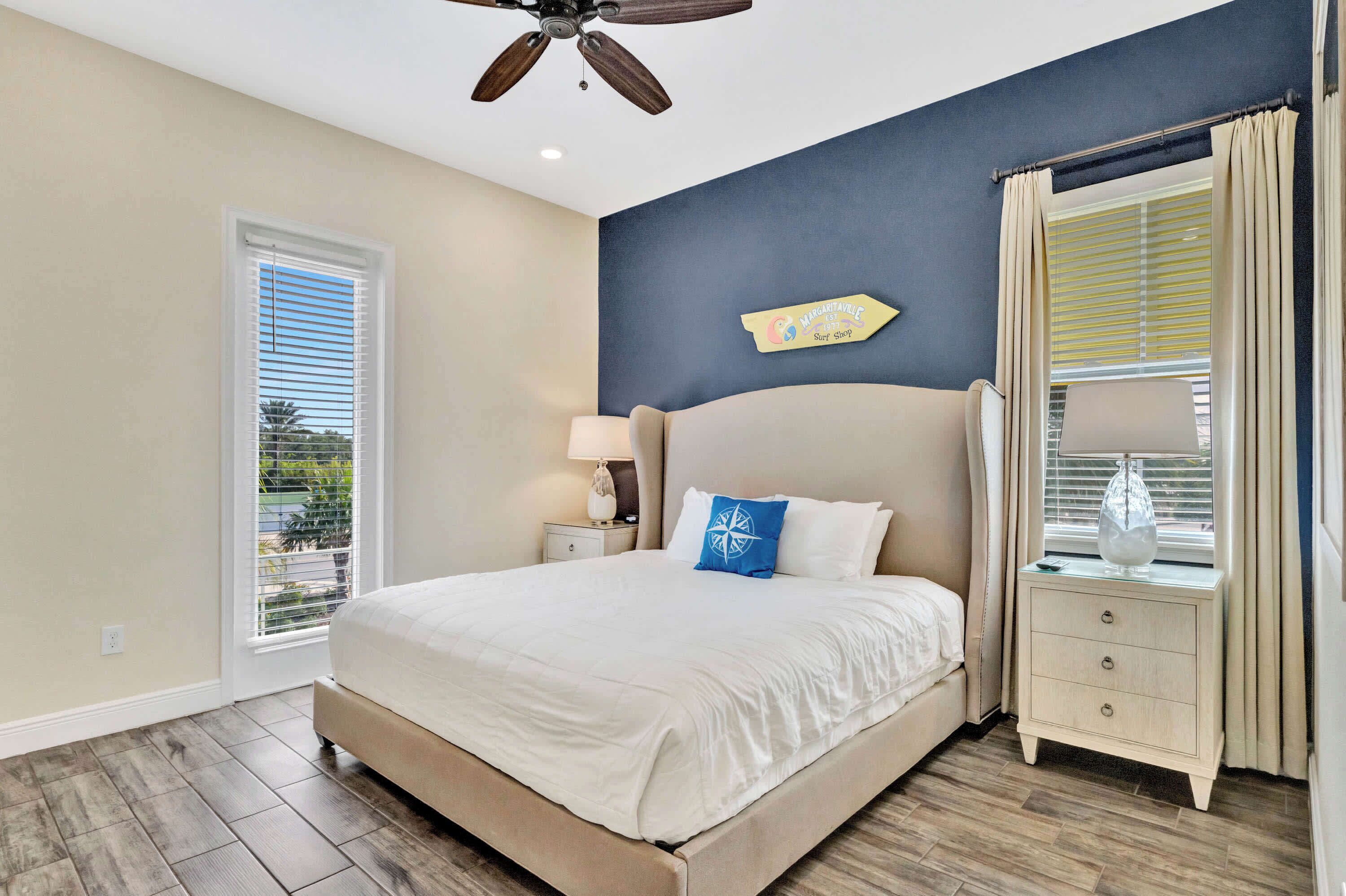 A bedroom with a large bed, a nightstand with a lamp, a ceiling fan, and a window with blinds.