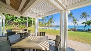 An outdoor patio with tables and chairs, overlooking a beach with palm trees and a clear sky. - Golfers Bliss (Kamuela)
