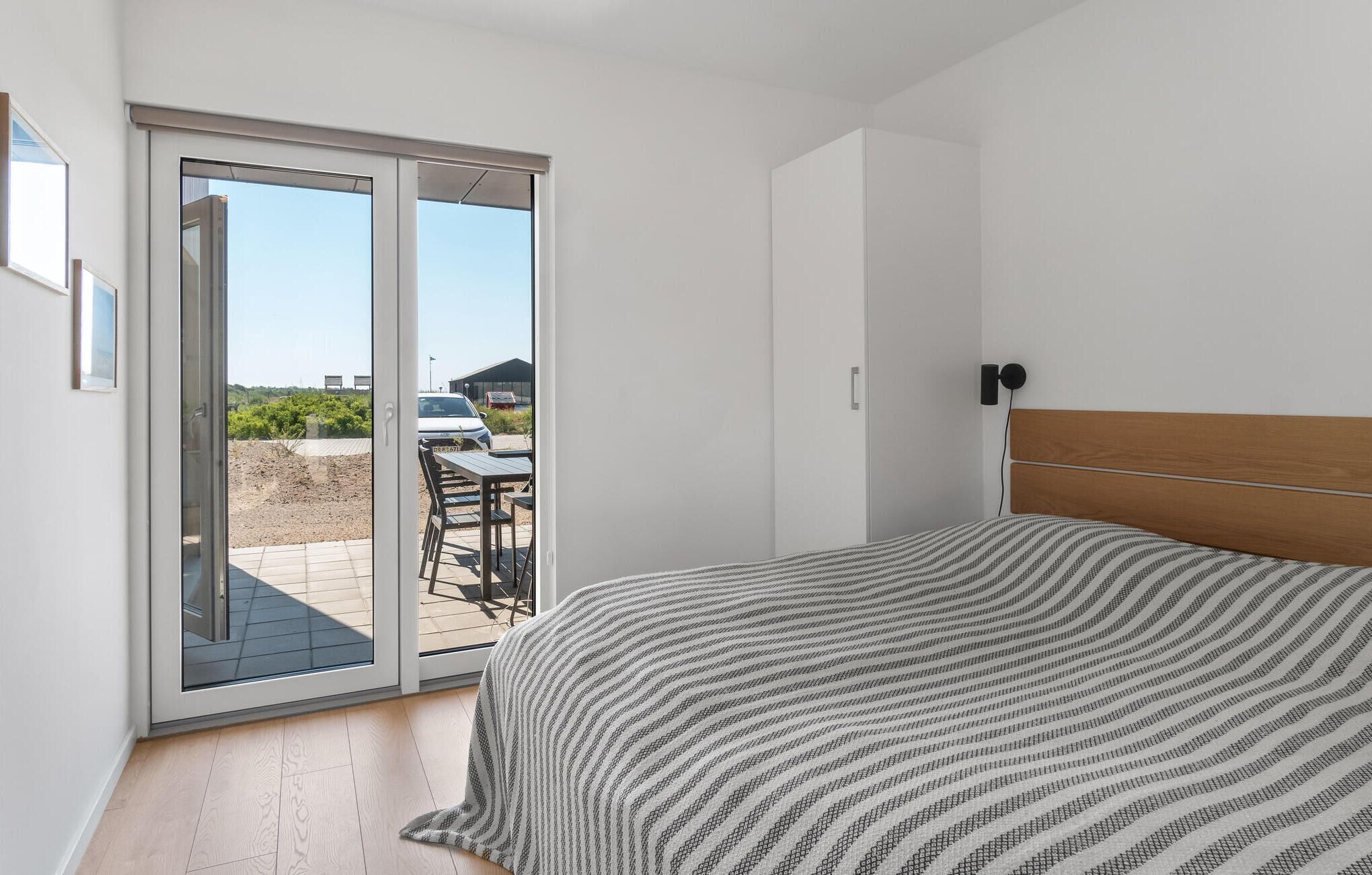 A bedroom with a bed, a sliding glass door leading to an outdoor area with a table and chairs, and a view of a car and greenery outside.
