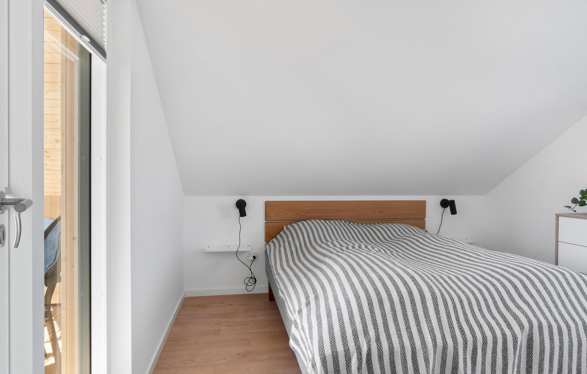 Une chambre avec un plafond en pente, une tête de lit en bois, un lit recouvert d’une couvre-lit à rayures et une petite table de chevet avec une lampe.