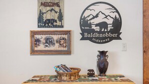 A wooden table with a basket of magazines, a framed picture, a decorative vase, and a welcome sign with a mountain scene.