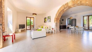 A spacious living area with a white sofa, a red table, and a dining set. An archway leads to a kitchen area with a refrigerator and a dining table.