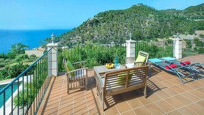 A terrace with a table and chairs, a pool, and a view of the sea and hills.